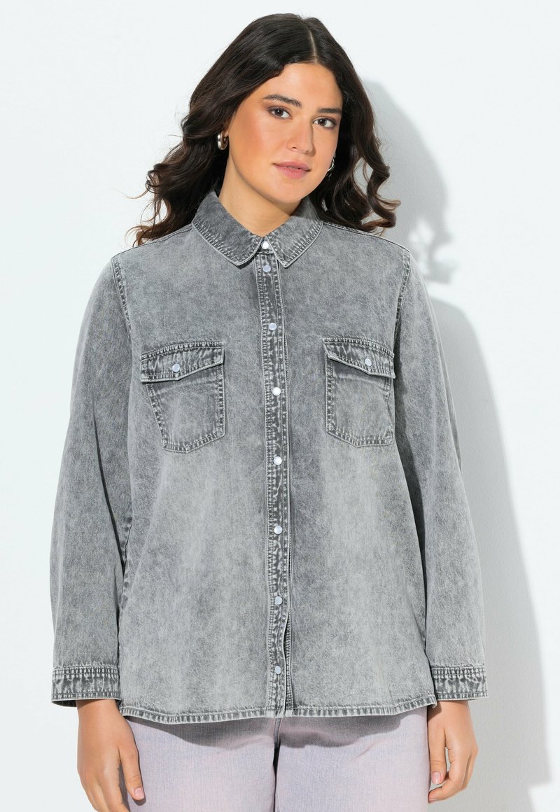 Camicia in denim grigio con colletto classico, chiusura a bottoni, due tasche sul petto e maniche lunghe. Indossata sopra pantaloni chiari.