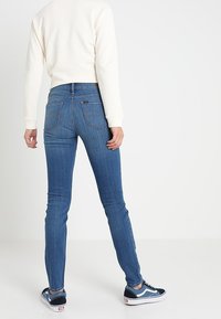 Pantalones vaqueros azul índigo de corte ajustado con cintura alta, cinco bolsillos y detalles de costuras sutiles. Combinados con zapatillas azul marino y blancas.