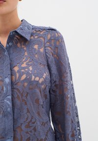 Cămiză din dantelă într-o nuanță de albastru deschis, cu modele florale, guler la gât și mâneci lungi cu detalii elaborate.