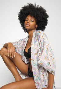 Kimono floral leve com cores pastel, apresentando um corte solto, mangas largas e padrões intrincados de flores e folhas. Bralette de renda preta por baixo.