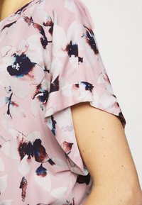 Détail de la manche d'un chemisier rose avec un grand imprimé floral en blanc, bleu marine et bordeaux sur le bras d'une personne, sur un fond clair.