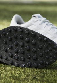 White athletic shoe with textured black sole featuring multiple hexagonal studs for traction. The upper has subtle stripes and a smooth finish.