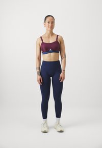 Reggiseno sportivo bordeaux con spalline regolabili, abbinato a leggings blu scuro a vita alta. Il modello si trova in un contesto neutro, indossando scarpe da ginnastica beige.