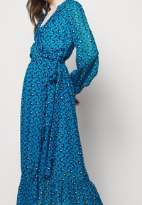 Vestido maxi floral azul hecho de una tela ligera, con mangas largas, diseño cruzado y un dobladillo con volantes con flores en contraste en blanco y negro.