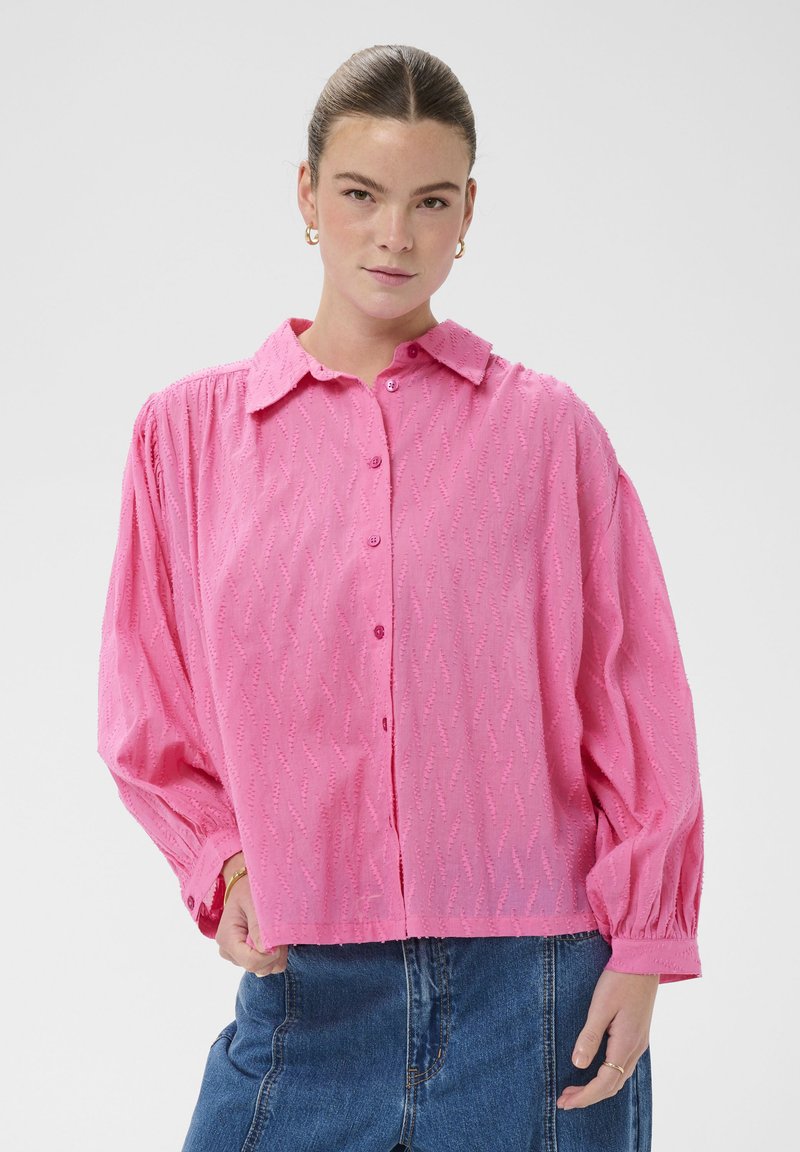 Chemise rose à boutons avec col et motif texturé. Elle présente des manches longues bouffantes avec des poignets élastiques et un ourlet droit.