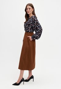 Kobieta stojąca bokiem, ubrana w wzorzystą bluzkę z długim rękawem, brązową spódnicę midi z kieszeniami i czarne buty na wysokim obcasie.