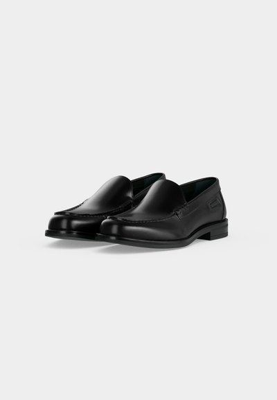 Ζευγάρι γυαλιστερά μαύρα δερμάτινα loafers slip-on με στρογγυλεμένες μύτες και χαμηλά στοιβαγμένα τακούνια σε απλό ανοιχτό φόντο.