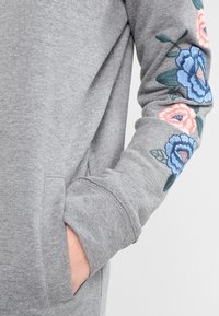 Grå sweatshirt med blomsterbroderi i blått och rosa på ärmen, med en ficka och mjukt, texturerat tyg.