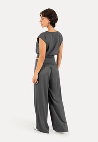 Femme aux cheveux courts portant un pantalon large gris foncé ample et un haut à manches courtes, debout et regardant sur le côté sur fond blanc.