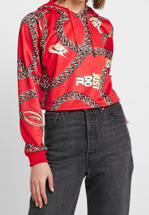 Sweat à capuche rouge Nike avec un imprimé chaîne noir et or, logo Nike blanc et texte "Just Do It", porté avec un jean noir taille haute.