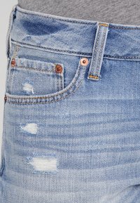 Jeans in denim blu chiaro con abrasioni vicino alla tasca anteriore e rivetti in rame, indossati con una maglia grigia in tessuto infilata dentro.