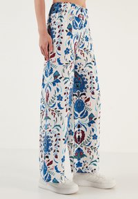 Wijdvallende broek van witte stof met een gedetailleerd bloemenpatroon in blauw en rood. Inclusief een elastische tailleband en gedragen met witte sneakers.