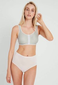 Ung kvinne iført en lys grå sports-BH med hvite detaljer og høyt midje beige truser, stående mot en ensfarget bakgrunn.