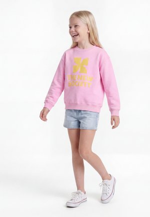 Lachend meisje met lang blond haar draagt een roze trui met gele tekst en logo, spijkershorts en witte sneakers, lopend naar links.