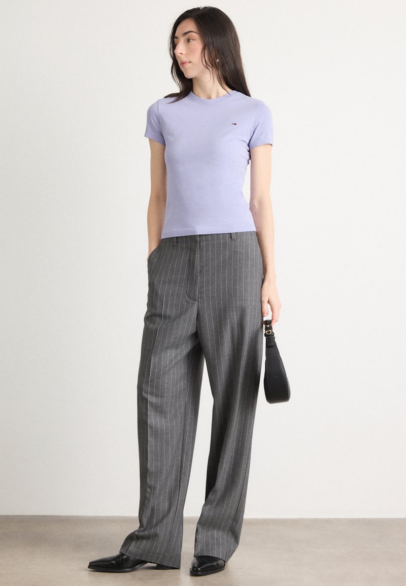 Lilla kortærmet t-shirt med et lille logo, parret med grå stribede bukser med vide ben. Modellen holder en sort håndtaske.