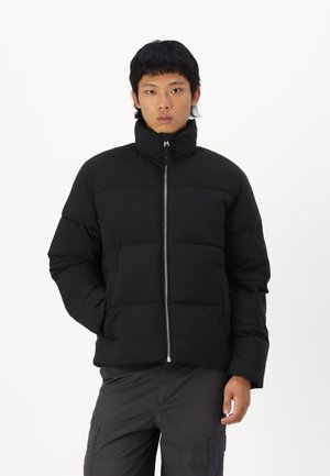 Giacca a vento nera con colletto alto, sezioni imbottite, chiusura con zip e una texture liscia, abbinata a pantaloni cargo scuri.