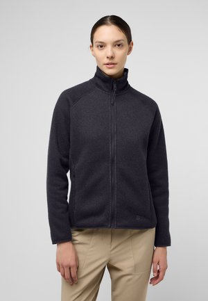 Veste en polaire gris foncé avec un col haut, fermeture éclair intégrale et texture côtelée. Elle présente des manches longues et des poches latérales, portée sur un pantalon beige.