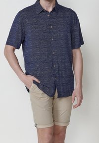 Homme portant une chemise bleu marine à manches courtes à motifs et boutonnée, ainsi qu’un short beige retroussé, une main dans la poche.