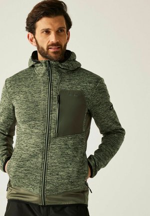 Mężczyzna ubrany w zieloną bluzę z kapturem zapinaną na zamek, wykonaną z teksturowanej tkaniny, z zamykaną kieszenią na piersi, z rękami w bocznych kieszeniach, patrzący lekko w prawo.