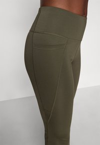 Højtaljede, olivengrønne leggings med et glat, strækbart stof. Har en sidelomme og kontrasterende syninger langs sømmene.