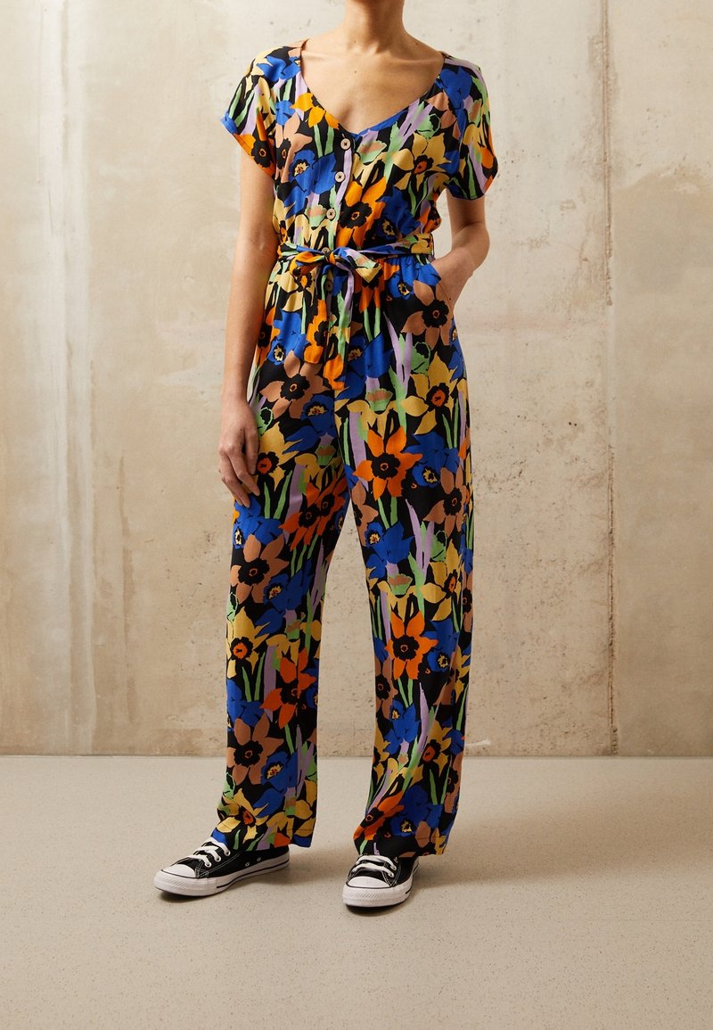 Femme portant une combinaison fleurie colorée avec des boutons, une taille nouée, des manches courtes et des baskets noires, debout contre un mur uni.