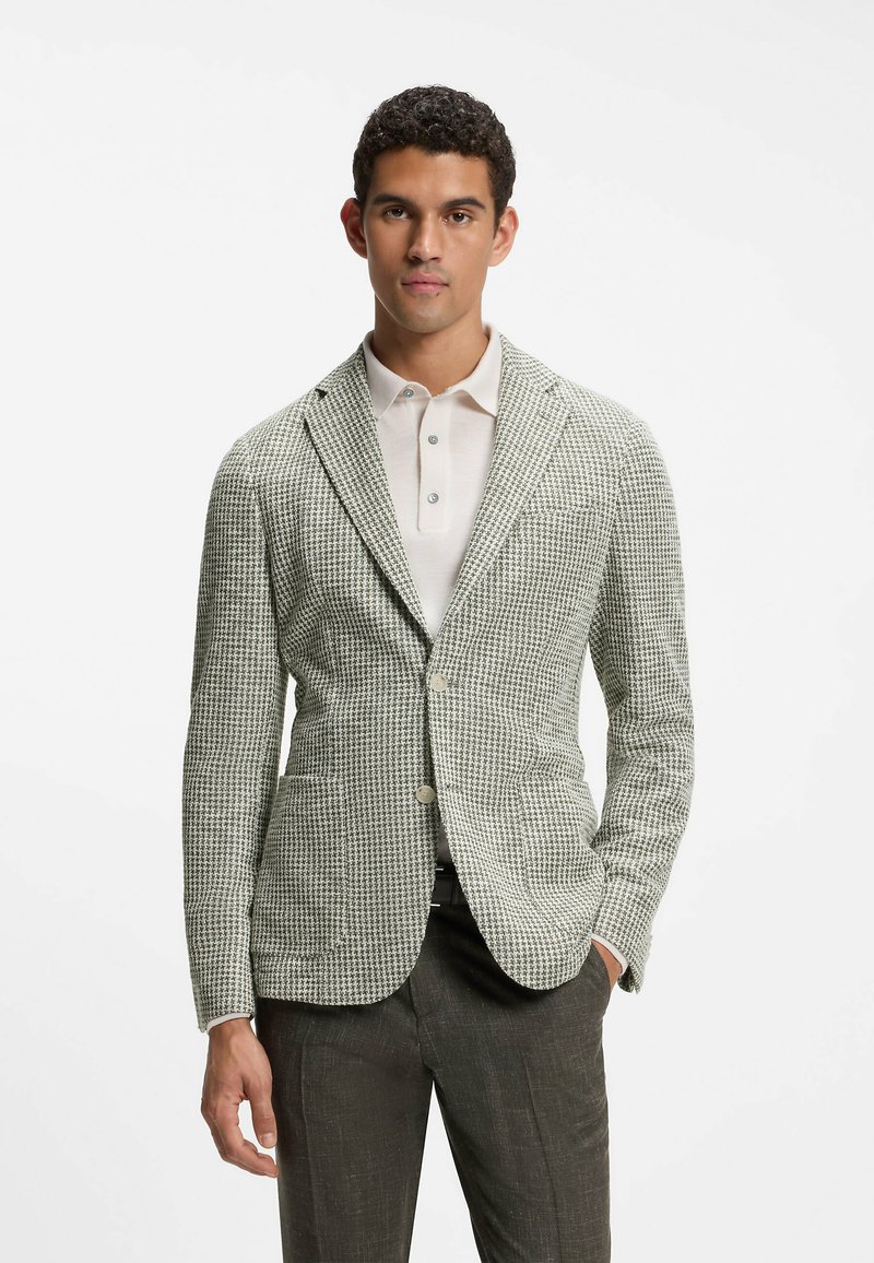 Homme portant un blazer pied-de-poule gris et blanc, un polo blanc et un pantalon foncé, debout avec une main dans la poche sur fond blanc.
