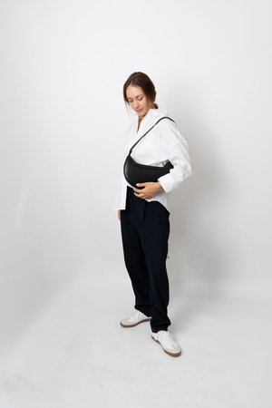 Mulher vestindo uma camisa branca e calças escuras com riscas verticais segurando uma bolsa preta em forma de meia-lua contra um fundo branco simples.