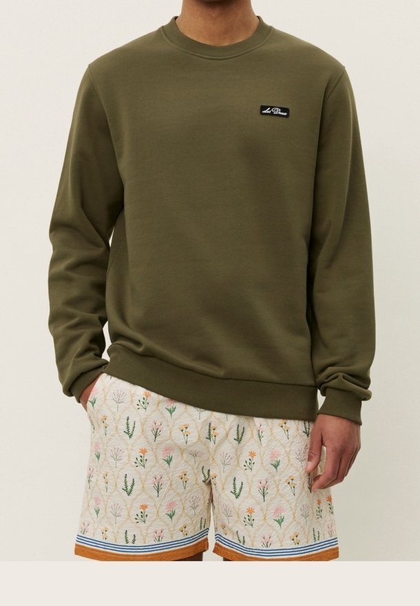 Homme portant un sweat-shirt vert olive et un short crème avec un motif floral multicolore et un ourlet rayé orange et bleu.