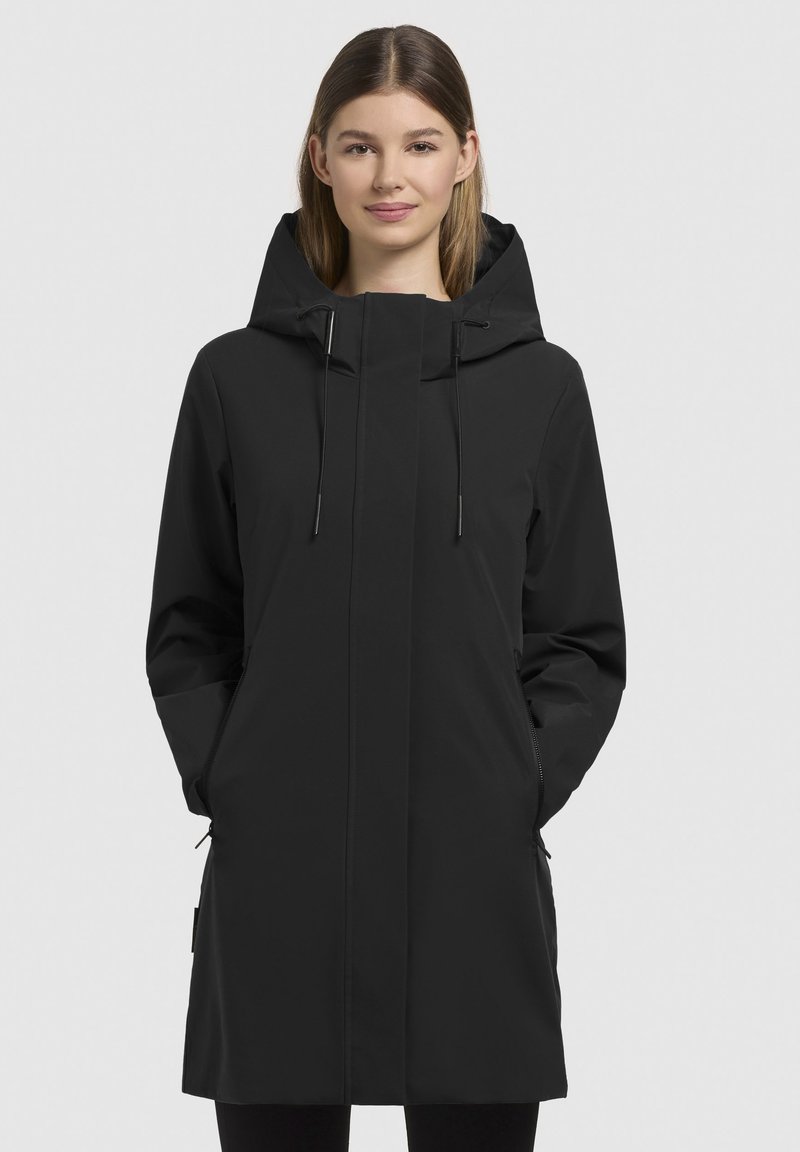 Manteau imperméable noir avec capuche, poches zippées et cordons de serrage réglables. Présente un design élégant et minimaliste ainsi qu'une texture lisse.