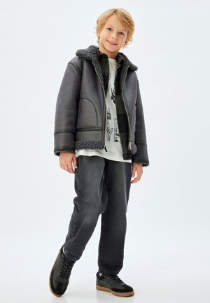 Chaqueta de piel de oveja gris forrada, con cierre de cremallera, combinada con jeans negros y zapatillas negras. El conjunto presenta un ajuste relajado y casual.