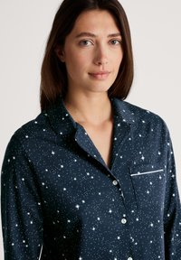 Donkerblauwe pyjamabovenkant met een sterrenpatroon, knoopsluiting en een borstzak met een witte rand. Zachte stof met een gladde textuur.