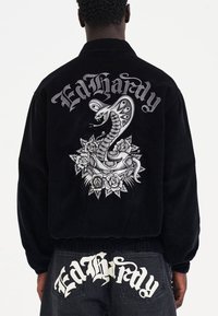 Musta korduroy-takki, jossa on selässä brodeerattu käärme- ja ruusukuvaus sekä "Ed Hardy" -teksti tyylitetyssä kirjasimessa.