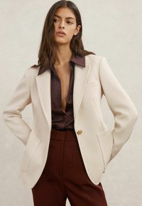 Beezhši bleiser struktureeritud disainiga, tekstureeritud kangas, sälguga kraed, kuldse nööbi detail, kombineeritud tumepruuni särgi ja lõhikuga pükstega.