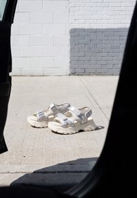 Des sandales blanches épaisses avec des semelles épaisses sont posées sur un trottoir en béton ensoleillé près d'un mur de briques blanc et gris.