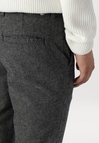 Pantalones de cuadros grises con una tela texturada y un brillo sutil. Presentan bolsillos y un corte ajustado, combinados con un suéter blanco de canalé.