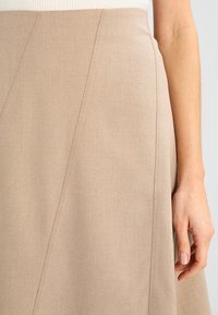 Jupe beige au design évasé, avec des coutures diagonales, une texture de tissu lisse et une taille ajustée. Main reposant sur le côté.