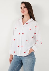 White button-up shirt made of lightweight fabric, featuring scattered red heart embroidery. Long sleeves with buttoned cuffs, casual fit.