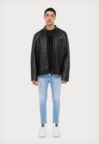 Schwarze Lederjacke mit Reißverschluss, ausgestattet mit einer Brusttasche und seitlichen Reißverschlüssen, kombiniert mit hellblauen Skinny-Jeans und weißen Sneakers.