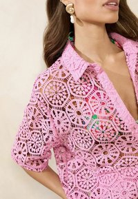 Rozā mežģīņu krekls ar sešstūrainiem ziedu rakstiem, ar apkakles izgriezumu un īsām piedurknēm, atklājot krāsainu iekšējo kārtu.