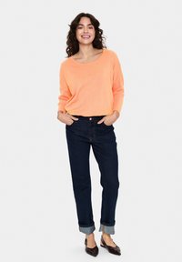 Oranger gestrickter Pullover mit weitem Halsausschnitt, kombiniert mit dunkelblauen Jeans mit Aufschlägen und braunen Flats. Model steht mit den Händen in den Taschen und lächelt.