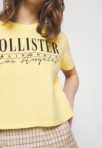 Žluté bavlněné tričko s výrazným černým potiskem "Hollister California Los Angeles". Krátké rukávy a volný střih.