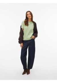 Grøn t-shirt med korte ærmer kombineret med en mørkebrun cardigan, løse blå cowboybukser og brune ankelstøvler, mod en hvid baggrund.