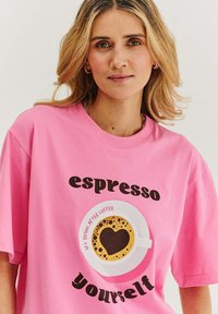 Růžové tričko s krátkým rukávem s kulatým potiskem šálku kávy s kávou ve tvaru srdce. Text zní "espresso yourself."