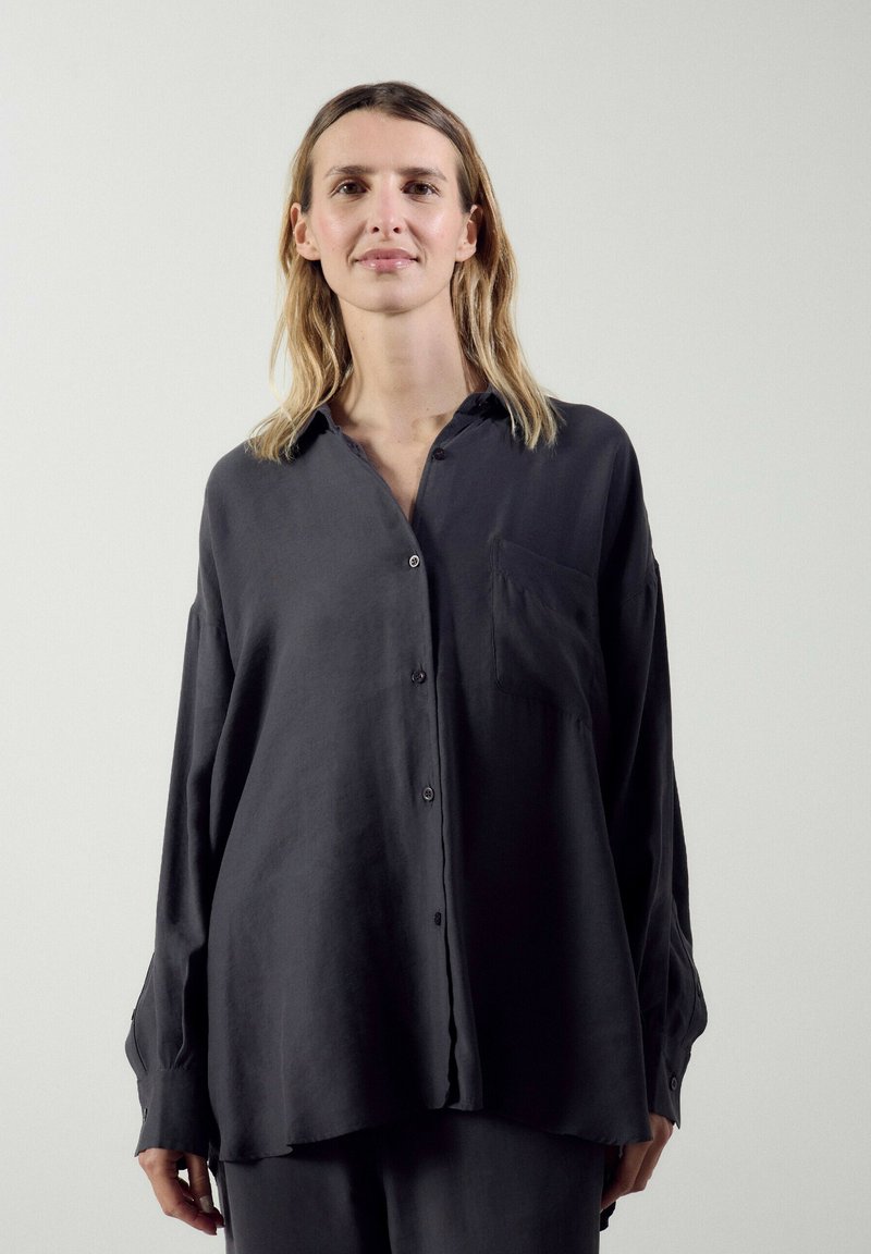 Femme aux cheveux blonds mi-longs portant une chemise noire ample à boutons sur un fond clair uni.