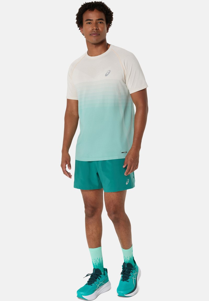 Mannen sportoutfit met een mintgroen en crèmekleurig T-shirt met korte mouwen in een gradient, teal shorts en teal hardloopschoenen met bijpassende sokken.