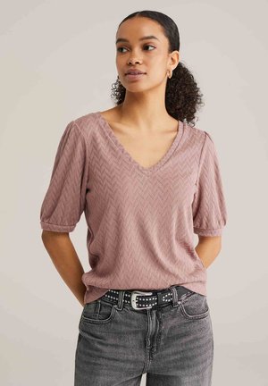 Kobieta z kręconymi włosami związanymi w kucyk, ubrana w fioletowy top z fakturą, dekoltem w serek i bufkami, czarny pasek z ćwiekami oraz szare dżinsy, z rękami za plecami.