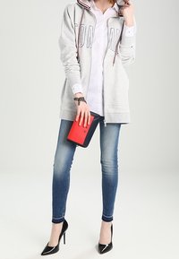Sudadera gris claro con cremallera, estampados en rojo y azul marino, camisa blanca de botones, jeans ajustados azules y tacones altos negros; sosteniendo un bolso de mano rojo y negro.