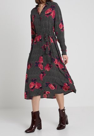 Femme portant une robe midi noire à imprimé floral rouge, manches longues et ceinture nouée à la taille, associée à des bottines à talons brillantes rouge foncé.