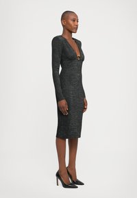 Woman in a fitted, long-sleeve black midi dress with deep V-neck and ring detail, wearing black pointed high heels, standing against white background.