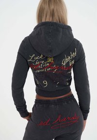 Žena v černé mikině a kalhotách s vyšitým žlutým a červeným textem, včetně "ed hardy," otočená zády na bílém pozadí.